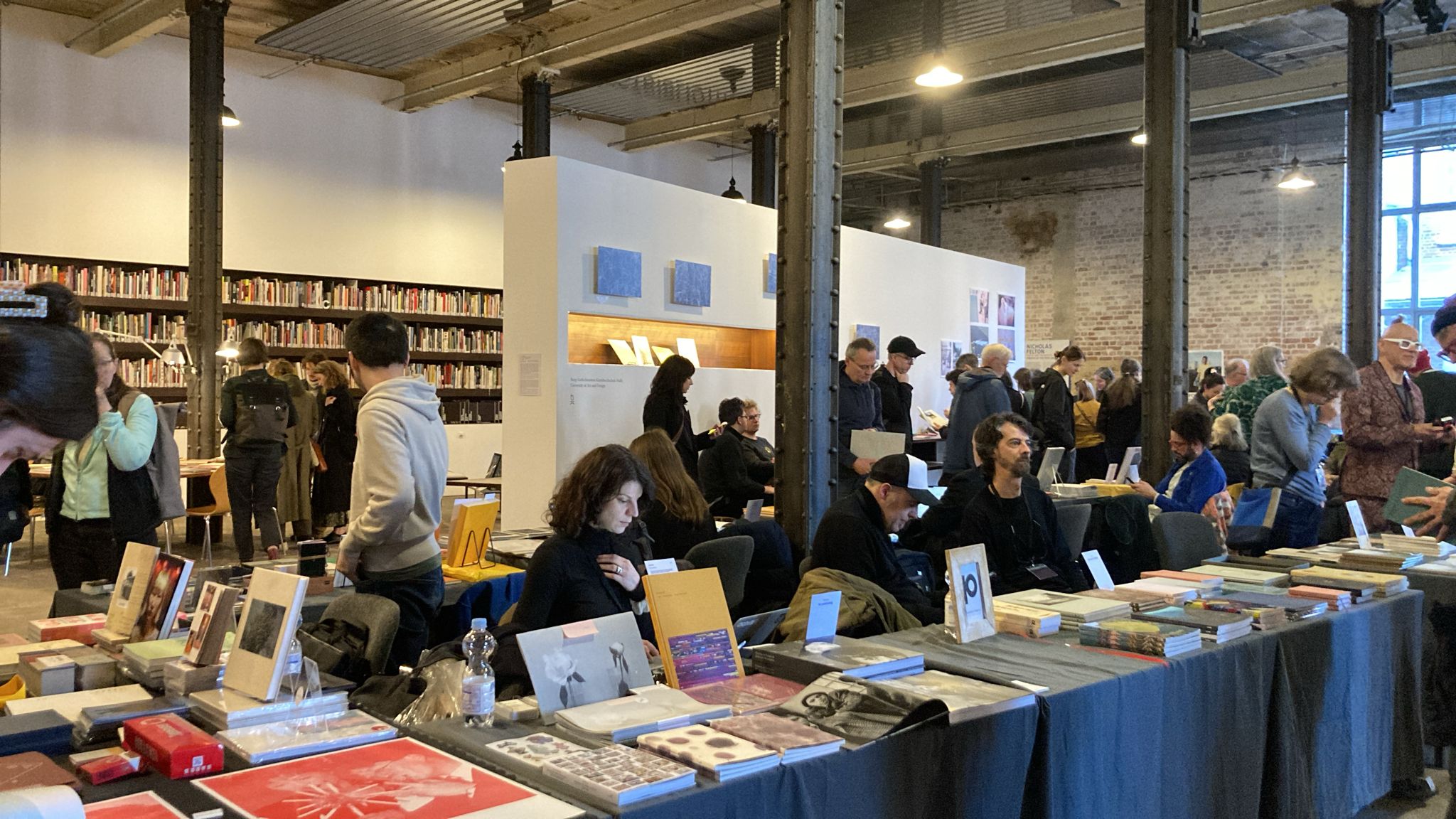 Leipzig Photobook Festival 2026 courtyard at Spinnerei with brick-red industrial buildings, cobblestone ground, and festival visitors gathered in afternoon sunlight